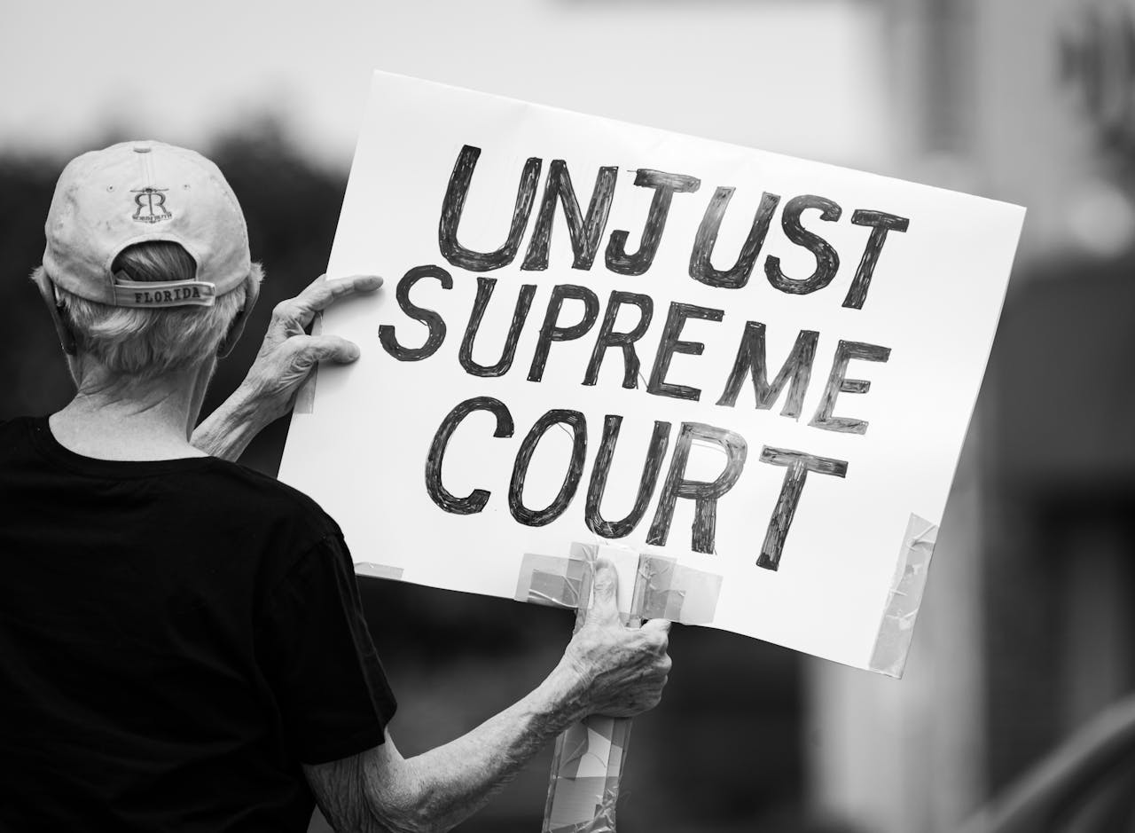 Services Black and white photo of a protester holding a sign saying 'Unjust Supreme Court' during a demonstration.